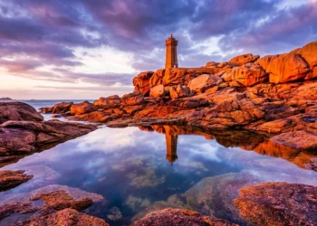 Paysage de la Côte de Granit Rose au coucher du soleil en hiver. Les rochers rougeoyants encadrent le phare de Men Ruz qui se reflète dans une mare d'eau de mer au premier plan.